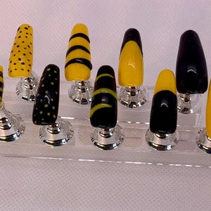 May include: A set of press-on nails in various designs, including yellow with black stripes, dots, and solid black and yellow tips. The nails are displayed on a clear, tiered stand with silver-colored bases.