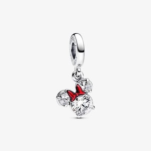 May include: A silver charm with a Minnie Mouse design. The charm features a large, clear, round crystal for the face, two smaller crystals for the ears, and a red bow. The charm is suspended from a silver loop.