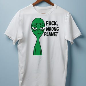 May include: White t-shirt featuring a green alien with a grumpy expression. The text "FUCK. WRONG PLANET" is printed in black. The shirt is hanging on a wooden hanger.
