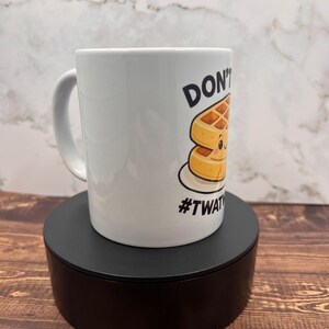 May include: White ceramic mug with a handle, featuring a cartoon waffle design and the text "DON'T" and "#TWAT". The mug is displayed on a black stand against a marble and wood background.