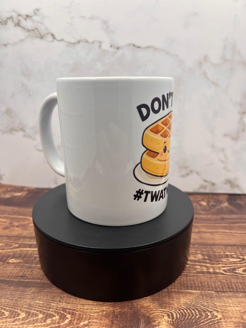 May include: White ceramic mug with a handle, featuring a cartoon waffle design and the text "DON'T" and "#TWAT". The mug is displayed on a black stand against a marble and wood background.