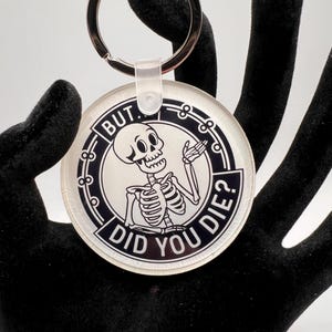 May include: A round, white, acrylic keychain with a black skeleton graphic and the text "BUT DID YOU DIE?" in a circle around the skeleton.