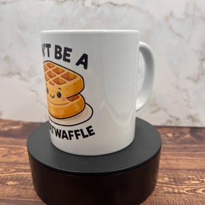 May include: White ceramic mug with the text "Don't be a Heatwaffle" and a cartoon waffle illustration. The waffle has a smiling face. The mug is on a black stand, set against a wooden surface.