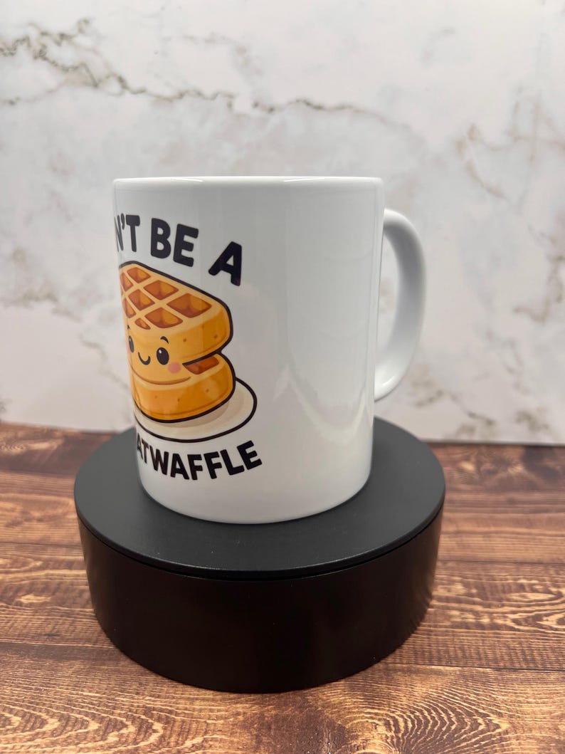 May include: White ceramic mug with the text "Don't be a Heatwaffle" and a cartoon waffle illustration. The waffle has a smiling face. The mug is on a black stand, set against a wooden surface.