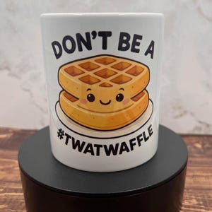 May include: White ceramic mug with the text "DON'T BE A #TWATWAFFLE" and a cartoon waffle design. The waffle is golden brown with a smiling face. The mug is on a black stand.