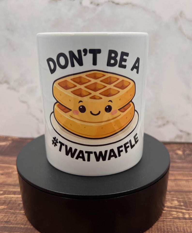 May include: White ceramic mug with the text "DON'T BE A #TWATWAFFLE" and a cartoon waffle design. The waffle is golden brown with a smiling face. The mug is on a black stand.