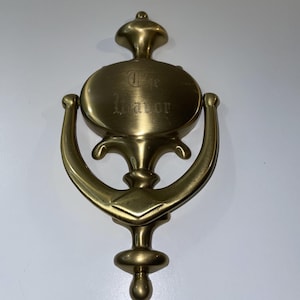 May include: A brass door knocker with a decorative design. The knocker features a circular plate with the words "The Manor" engraved on it. The knocker has a curved handle and a decorative base. The overall design is ornate and classic.