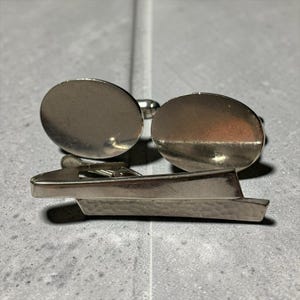 May include: Silver-toned cufflinks and tie clip set. The cufflinks have an oval design. The tie clip is shaped like a boat. The set is made of a shiny metal and is displayed on a gray surface.