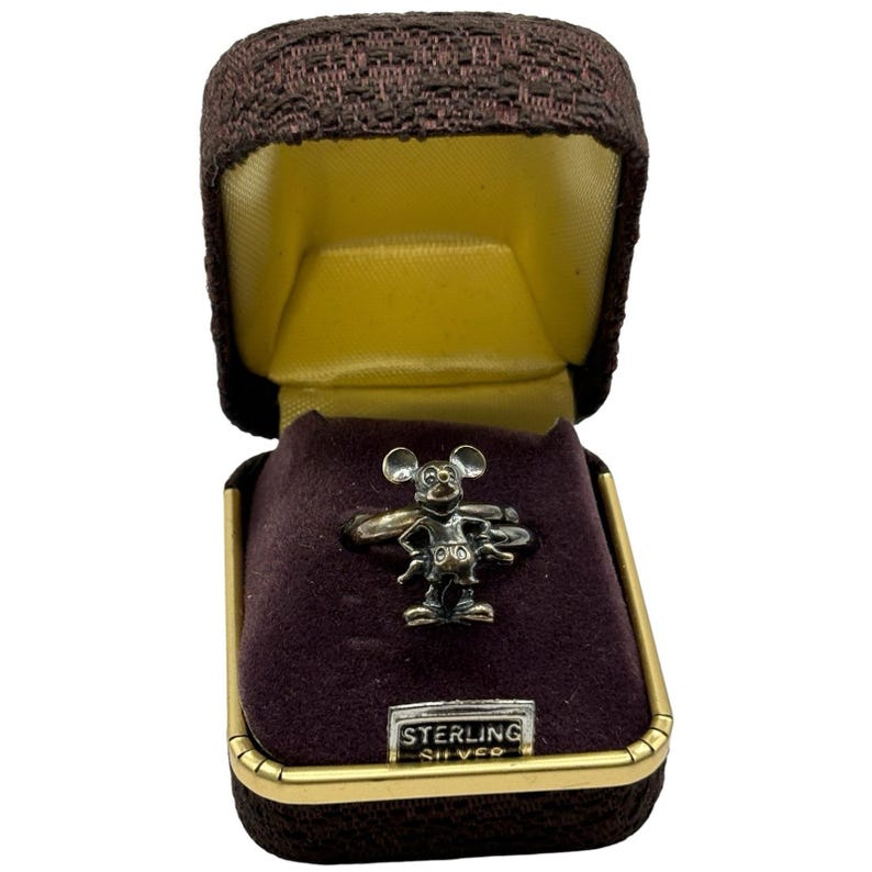May include: A silver Mickey Mouse ring in a brown velvet box with a gold trim. The box has a label that reads "Sterling Silver".