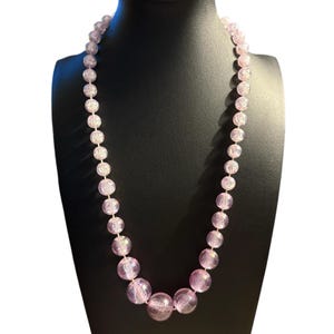 May include: A pink beaded necklace with varying sizes of round beads. The beads have a crackled, textured appearance and are strung together. The necklace is displayed on a black mannequin bust.