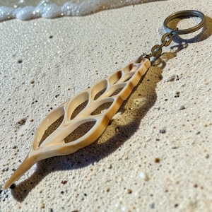 May include: A natural seashell keychain with a light tan color and a unique, openwork design. The keychain is attached to a metal ring and chain, resting on a sandy beach, capturing a coastal theme.