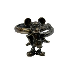 May include: A silver-toned ring featuring a Mickey Mouse figurine. The ring is a twisted band with a textured finish.