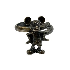 May include: A silver ring featuring a Mickey Mouse figurine. The ring is twisted and the figurine is detailed with black eyes and a red nose.