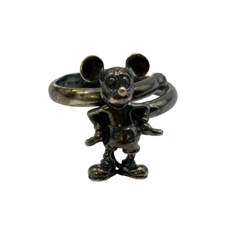 May include: A silver ring featuring a Mickey Mouse figurine. The ring is twisted and the figurine is detailed with black eyes and a red nose.