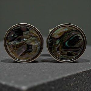 May include: A pair of round cufflinks with iridescent abalone shell inlays. The cufflinks have a silver-colored metal border and a dark background. The abalone shell displays a mix of colors, including green, blue, and purple.