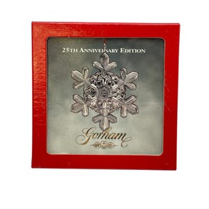 May include: A silver snowflake ornament inside a red box. The ornament is detailed with intricate patterns and the box reads "25th Anniversary Edition" and "Gorham". A festive holiday decoration.