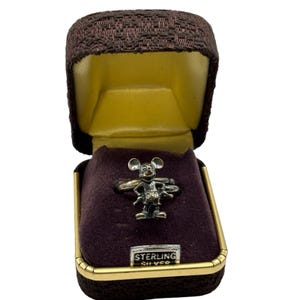 May include: A sterling silver Mickey Mouse ring in a burgundy velvet ring box with a gold trim. The box has a yellow interior and the words "STERLING SILVER" are printed on the bottom.