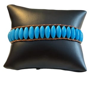 May include: A wrap bracelet featuring bright turquoise oval beads strung on a brown leather cord. The bracelet is displayed on a black surface, showcasing the vibrant color and design of the jewelry.