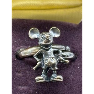 May include: A silver ring featuring a Mickey Mouse figurine on top. The ring is displayed on a dark purple velvet surface.