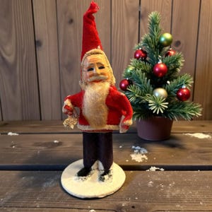 May include: A vintage Santa Claus figurine with a red coat and hat, white beard, and black boots. The figurine is standing on a white base. A small Christmas tree with red and gold ornaments is in the background.