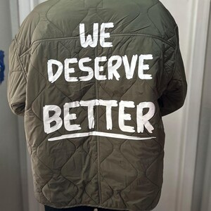 May include: An olive green quilted jacket featuring the white text "WE DESERVE BETTER" on the back. The jacket has a hood and a diamond pattern. The text is in a bold, sans-serif font.