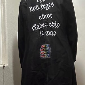 May include: Black long coat with white gothic lettering on the back. The text reads "amor liberabit vos non reges amor clades odio te amo." A colorful patch with the words "Queer Witches Against Fascism" is also visible.