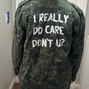 Może przedstawiać: Zielona kurtka moro z napisem "I REALLY DO CARE DON'T U?" w białych literach na plecach. Kurtka ma talię ze sznurkiem i pagony.