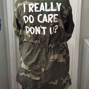 Może przedstawiać: Kurtka moro z napisem "I REALLY DO CARE DON'T U?" w białych literach. Kurtka posiada talię ze sznurkiem i klasyczny wzór inspirowany wojskiem w odcieniach zieleni, brązu i beżu.