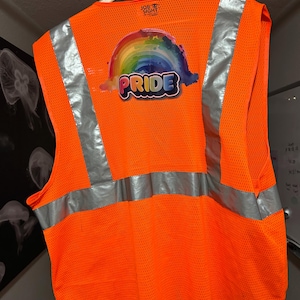 May include: Bright orange safety vest with reflective silver stripes and a rainbow Pride graphic. The vest has the word "PRIDE" in a colorful font and the words "JOB SIGHT" at the top. The vest is hanging on a wooden hanger.