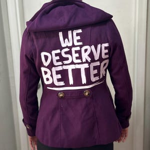 May include: A dark purple jacket with a hood, featuring the white text "WE DESERVE BETTER" on the back. The jacket has two rows of buttons and a tailored design. The hood is up, and the fabric looks soft.