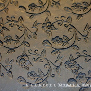 May include: A close-up of a dark gray textured surface with a repeating pattern of stylized flowers and vines. The surface is a stamp for creating decorative impressions.