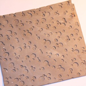 May include: A square, light brown clay tile with a repeating pattern of small, raised circles.