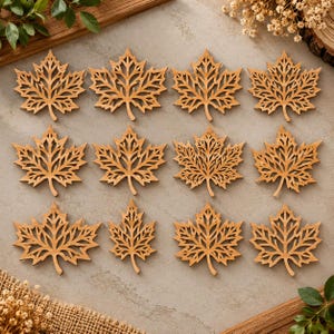 May include: A set of twelve light brown wooden maple leaf cutouts arranged in a grid. Each leaf features a detailed, intricate laser-cut filigree pattern with sharp points and a central stem, showcasing a natural wood grain texture throughout.