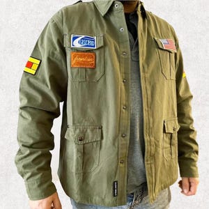 Könnte beinhalten: Olivgrünes Hemd mit mehreren Taschen und Aufnähern. Zu den Aufnähern gehören ein blau-weißes Logo, ein brauner rechteckiger Aufnäher, eine kleine amerikanische Flagge und ein gelb-roter Aufnäher. Das Hemd hat einen klassischen Kragen und lange Ärmel.