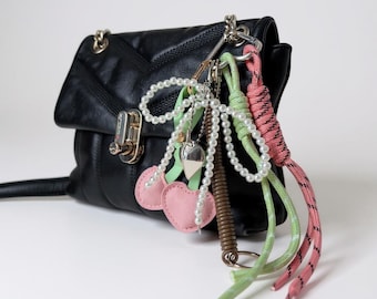 May include: Black leather handbag with a silver chain strap and clasp. The bag is adorned with a collection of charms, including pink felt cherries, pearl strands, and colorful rope tassels. A stylish accessory.