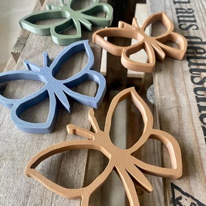 Könnte beinhalten: Vier Holzschmetterlingsformen in verschiedenen Farben: grün, braun, blau und beige. Die Schmetterlinge sind auf einer Holzoberfläche angeordnet.