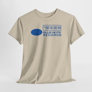 Puede incluir: Una camiseta beige con el texto azul "THE FINEST JAZZ SINCE 1939" en un rectángulo, y un óvalo azul. Debajo, las palabras "BLUE NOTE RECORDS" están impresas en azul.