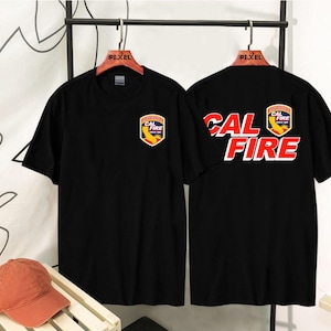 May include: Black t-shirts with the text "CAL FIRE" in red and white, and a badge logo. The front of the shirt has a smaller badge. A burnt orange baseball cap rests on a white slatted surface.