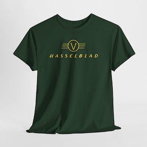 Può includere: T-shirt verde scuro con logo Hasselblad giallo. Il logo presenta un cerchio con una 'V' all'interno, affiancato da linee orizzontali, e il nome del marchio sottostante in un carattere giallo coordinato. La maglietta ha maniche corte e girocollo.