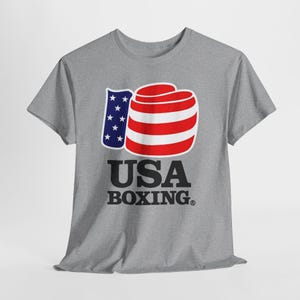 May include: A gray t-shirt featuring a boxing glove design in red and white stripes, with a blue section containing white stars. The text "USA BOXING" is printed below the graphic in black.