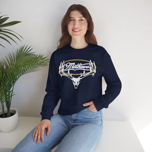 May include: Navy blue sweatshirt featuring a white and gold design with the word "Mathews" and a deer skull graphic. The long-sleeved crew neck top is made of a soft material and is suitable for casual wear.