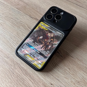 Op de afbeelding: Een zwarte smartphonehoes met een transparant achterpaneel waarin een Umbreon en Darkrai GX Tag Team Pokémon-verzamelkaart is geplaatst. De hoes heeft een verhoogde uitsparing voor de camera en een strak, beschermend frame.