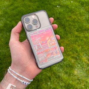 May include: Clear phone case with a Pokemon card featuring a pink and white illustration of Mew EX. The phone case has a black border and is held against a green grassy background. The card shows a colorful scene with buildings and characters.