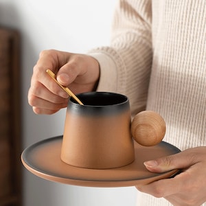 May include: A brown and black ceramic coffee cup with a wooden handle and matching saucer. A small gold spoon rests inside the cup. The cup and saucer are held by a person.