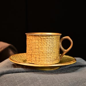 以下が含まれることがあります： 黄色のセラミックティーカップとソーサーのセット。カップは丸みを帯びたハンドル付きで、お揃いのソーサーの上に置かれています。セットは格子状のパターンと濃い茶色の縁があります。背景は暗いです。