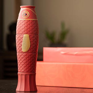 May include: A red fish-shaped container with a gold accent on the side and top. The container is made of four stackable pieces. The fish has a textured, scale-like pattern. The container is next to two red boxes.