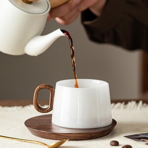May include: A white ceramic mug with a wooden handle and a matching wooden saucer. Dark liquid is being poured from a white teapot into the mug. The mug is sitting on a wooden saucer, with a gold spoon and coffee beans nearby.