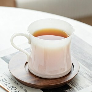 May include: A white ceramic teacup filled with tea, on a round wooden coaster. The cup has a curved handle and flared rim. The tea is amber, and the coaster is dark brown. The cup and coaster are on a table.