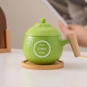 Könnte beinhalten: Eine hellgrüne Keramiktasse mit Deckel, Holzgriff und Untersetzer. Auf der Tasse befindet sich ein weißer runder Aufkleber mit dem Text "Your LOGO Here". Die Tasse ist wie eine Limette geformt.
