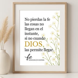 Puede incluir: Una impresión enmarcada con un marco de madera clara. La impresión presenta texto en español con la palabra "DIOS" en dorado. Flores blancas y tallos verdes están en el lado derecho. Un jarrón con plantas secas está a la izquierda.